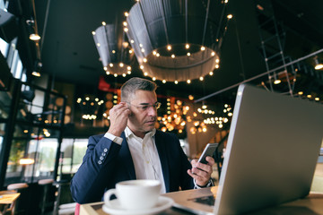 Middle-aged Caucasian man with serious face dressed smart casual using smart phone for call while sitting in cafe. In the desk laptop and coffee.