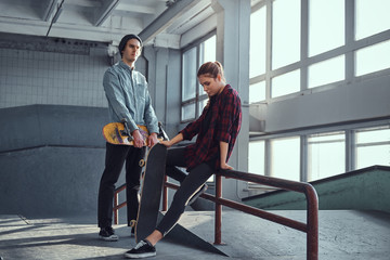 Attractive youth couple with skateboards next to a grind rail in skatepark indoors.