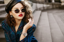 Outdoor Fashion Portrait Of Young Beautiful Fashionable Girl Wearing Trendy Sunglasses, Wrist Watch, Earrings, Leather Beret, Blue Checkered Dress, Posing In Street, On Stairs. Copy, Empty Space
