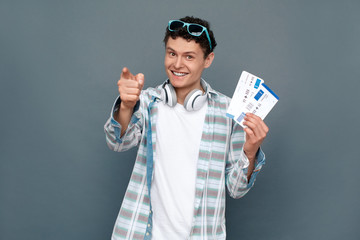 Man isolated on gray wall tourism concept standing holding two tickets pointing at camera happy