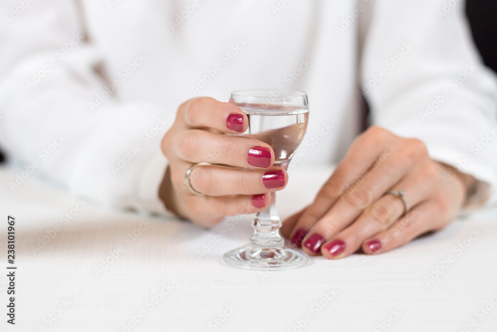 Kobieca dłoń z umalowanymi na czerwono paznokciami trzyma kieliszek wypełniony wódką. - obrazy, fototapety, plakaty 
