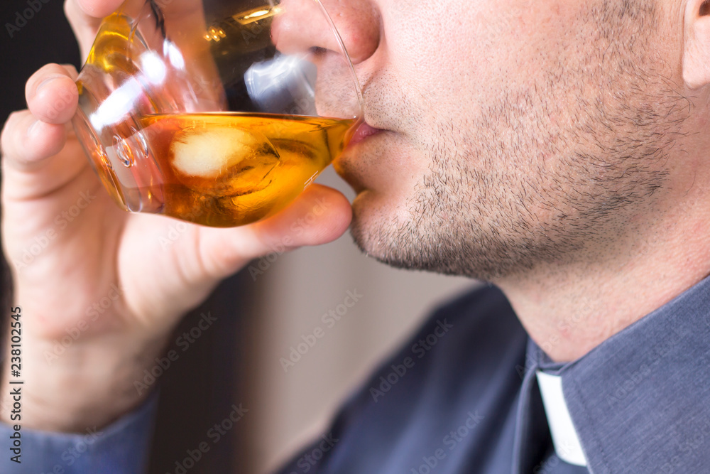 Ksiądz w koloratce i szarej koszuli pije whisky z lodem ze szklanki. - obrazy, fototapety, plakaty 
