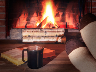 Wall Mural - Cup of tea, book, women's feet in warm socks on a wooden table opposite a burning fireplace