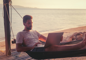 Young attractive guy a man with a beard in a hammock with a laptop on the background of the sea and sunset, distant work, freelancer, blogger, vacation and travel