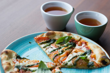 Sliced Pizza Margarita or Margherita lies on a blue plate against a wooden table and two cup of tea background. Pizza with Tomatoes, Fresh Basil, Mozzarella Cheese, Pepper and Spices close up. Traditi