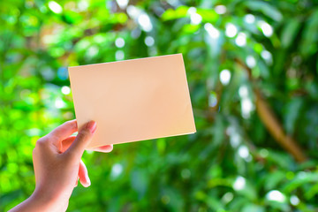 A woman's hand holding a blank empty yellow color paper wth green nature background.