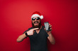 © Vulp - Cheerful happy bearded man wearing santa claus hat pointing at smartphone over red background