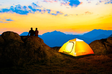 Wall Mural - young couple sitting on the rock near illuminated tent and watching the beautiful evening mountain v