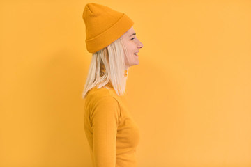 Wall Mural - Profile portrait of happy blonde hair woman wears yellow clothes, hat, smiles positively, isolated over yellow background. Copy space for advertisement or promotion. Caucasian female stands sideways