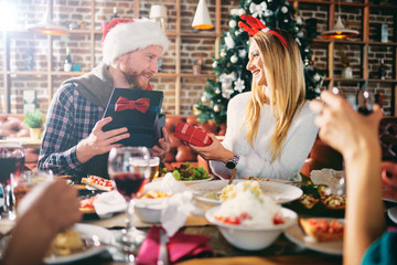 Wall Mural - Friends giving gifts to each other while sitting at table. In background Christmas tree. Christmas holidays concept.