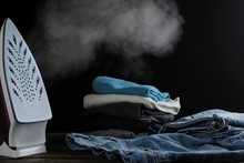 Lilac Iron Releases Steam And A Stack Of Clothes On A Black Background. Ironing Clothes. Household Electrical Appliances.