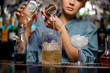 Female bartender pouring to the measuring cup with ice cubes an alcoholic drink from steel jigger