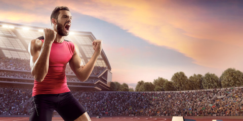 Wall Mural - Happy male athlete. Man sprinter in sport clothes emotionally rejoices victory at the running track in professional stadium