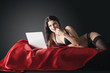 © Vitte Yevhen - Sexy young brunette woman in black sensual lingerie and in stockings using laptop on red bed in studio.