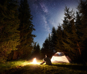 Wall Mural - Camping site at night. Tourist tent on forest clearing and male hiker sitting in front of burning campfire under night blue starry sky with Milky way on pine trees background. Outdoor activity concept