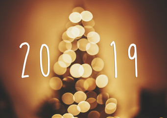 Wall Mural - 2019 text on christmas tree golden lights in festive room, blur bokeh of  glowing decoration on tree branches. Happy New Year. Season's greetings card. Merry Christmas