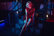 Beautiful glam blond woman with provocative make up wearing red short fitted sequin dress sitting on the stairs in the night club in colourful neon lights. Loft interior. Text space