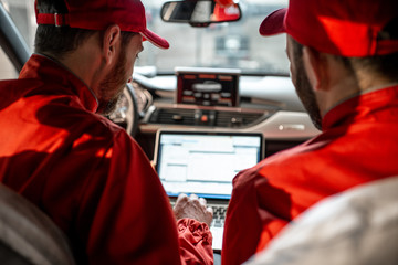 Wall Mural - Two male auto mechanics in red uniform diagnosing car with computer sitting on the seats indoors. Back view