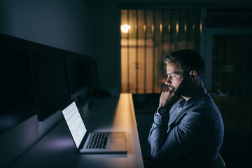 Young concerned bearded Caucasian businessman looking at laptop and finding a solution for the problem while sitting late at night in the office.