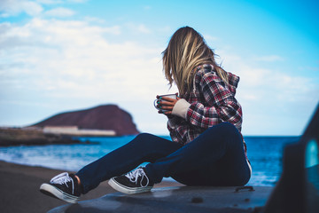 Rear back view of beautiful blonde hair woman sitting on the car and looking at the horizon with blue ocean and sky in background - enjoy life and travel lifestyle for digital nomad millennial people