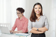 © DragonImages - Pretty young ethnic woman standing with arms crossed smiling at camera with coworker on background