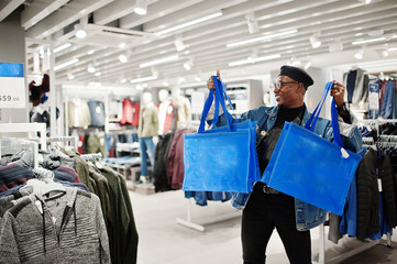 Wall Mural - Stylish casual african american man at jeans jacket and black beret with fanny pack or waist bag holding blue shopping bags at clothes store.
