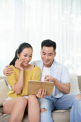 Wall Mural - Embracing Asian couple sitting on sofa at home sharing tablet and having fun 