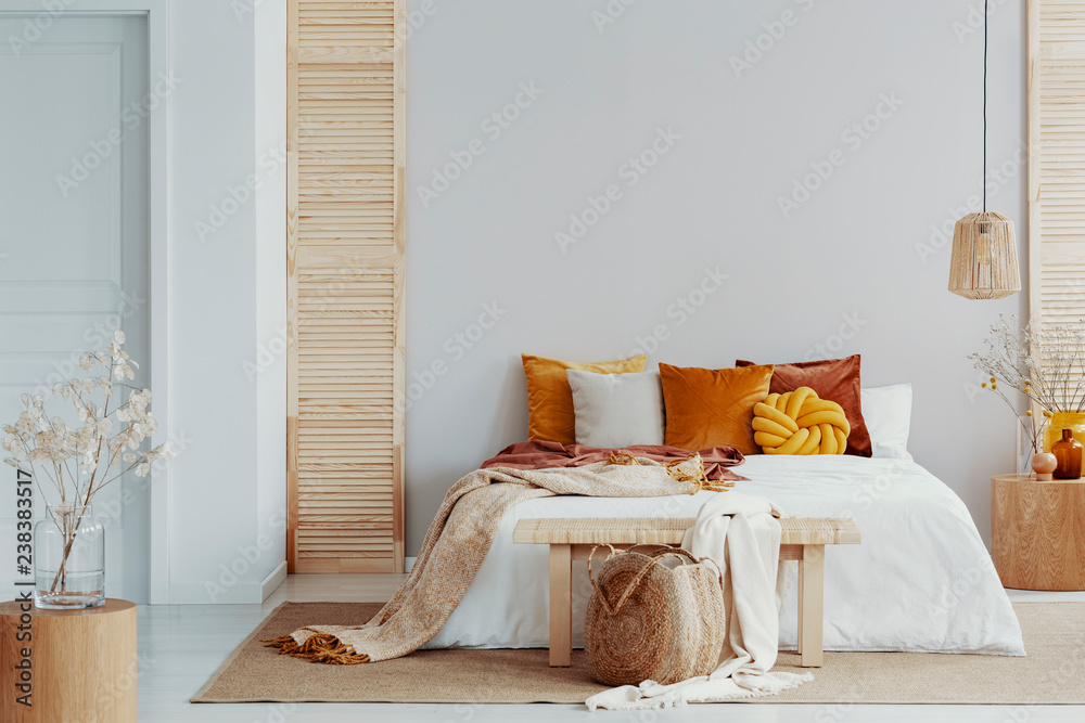 Brown and orange pillows on white bed in natural bedroom interior with wicker lamp and wooden bedside table with vase - obrazy, fototapety, plakaty 