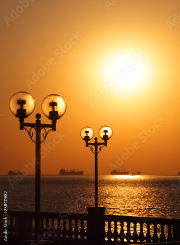 Fotografía Scenic view of waterfront with lanterns backlit by setting sun and with ships in the roadstead