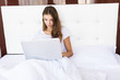 © dianagrytsku - Happy young brunette woman using laptop while sitting on bed in the morning
