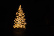 Silhouette of blurry Christmas tree illuminated and decorated with golden fairy lights in dark night creating abstract bokeh circles and glowing dots, image for holiday card with text copy space