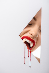 Wall Mural - cropped shot of woman with vampire teeth and blood biting through hole on white