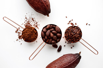 Rose gold measuring cups of cocoa beans, cacao nips, cocoa powder and cocoa pods on a white background, flat lay healthy food concept