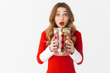 Sticker - Portrait of surprised woman 20s wearing Santa Claus red costume smiling and holding Christmas tree decorations, isolated over white background