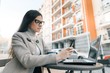 © Valerii Honcharuk - Young beautiful girl student with glasses in warm coat sitting in an outdoor cafe with laptop computer with phone cup of hot drink, city street background
