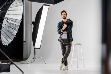 Wall Mural - full length view of confident young photographer holding professional camera and looking at camera in photo studio