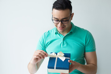 Wall Mural - Curious man wearing polo t-shirt and unwrapping gift box. Man untying ribbon bow. Surprise concept. Isolated front view on white background.