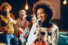 Close Up Of Mixed Race Woman Singing. In Background Band Playing Instruments. Home Studio Interior.