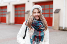 Portrait Of A Cheerful Positive Young Woman With A Sweet Smile With A Warm Stylish Checkered Scarf And White Fashionable Jacket Against The Background Of A Building With Red Doors. The Girl Smiles.