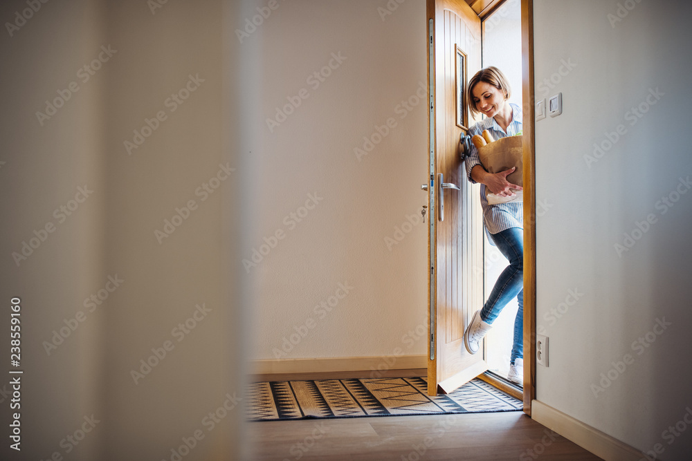 A young woman with groceries in paper shopping bag walking in through front door. - obrazy, fototapety, plakaty 