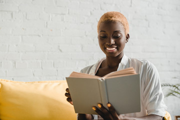 cheerful african american woman with short hair reading book on yellow sofa