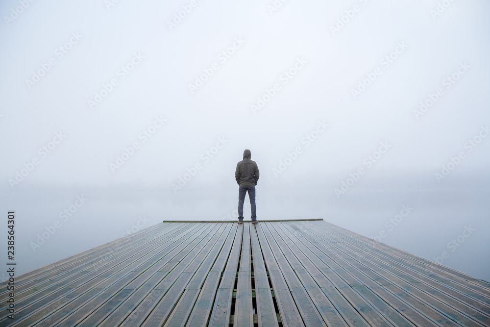 Young man standing alone on edge of footbridge and staring at lake. Mist over water. Foggy air. Early chilly morning in autumn. Beautiful freedom moment and peaceful atmosphere in nature. Back view. - obrazy, fototapety, plakaty 