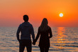 Man and woman holding each other hands. Young couple standing and staring at small waves of sea and orange sunset. Silhouettes on beach. Peaceful atmosphere in summer evening. Back view.