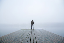 Young Man Standing Alone On Edge Of Footbridge And Staring At Lake. Mist Over Water. Foggy Air. Early Chilly Morning In Autumn. Beautiful Freedom Moment And Peaceful Atmosphere In Nature. Back View.