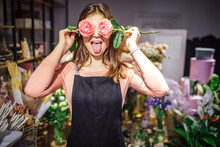 Funny And Positive Young Female Florist Cover Eyes With Roses And Show Tongue. She Has Fun. Plants And Flowers Stand In Vases Behind.