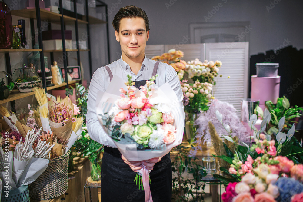Handsome young male florist stand and look on camera. He hold bouquet of flowers in hands. Guy stand inside. He cheerful. - obrazy, fototapety, plakaty 