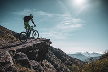 Mountain Biker Riding Up A Large Rock Deep In The Alps With Sun And Mountain Layers Behind
