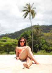 Wall Mural - Young woman in bikini and sunglasses, sitting on the beach sand, smiling, palms behind her.