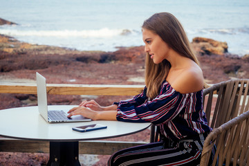 beautiful and young business woman in a stylish striped blouse with open shoulders sitting at the table with a laptop on terrace of the summer restaurant on the ocean.freelancer girl working online