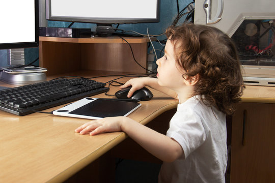 Little 2 3 year old baby girl in white clothers draws at the home computer in graphics drawing tablet. two monitors. The child is holding a mouse. Mock up vith copy space for text
